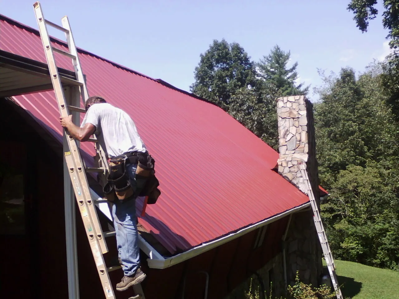 Roofer on a ladder beside a red metal roof and stone chimney — Soffit & Fascia Repair by Roofing Brothers in Copper Hill